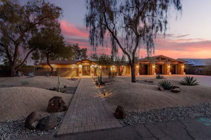 Sunset-lit Southwestern desert ranch home glowing with warm entry lights, a paver walkway through xeriscape landscaping of cacti and agave, and a three-car garage.