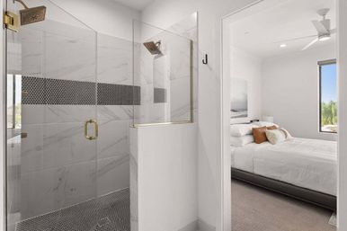 Bright contemporary ensuite with glass walk-in shower, gold fixtures, marble-look tiles and black penny-tile accent, opening into a white-bedded bedroom with throw pillows and a window view.