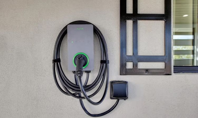 Wall-mounted home electric vehicle (EV) charging station with coiled black cable and plug on a beige stucco exterior wall beside a modern window frame.