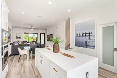 Bright modern open-concept kitchen and living area with white island and gold hardware, wood-look floors, dining table, sitting area and sliding glass doors to a green backyard.