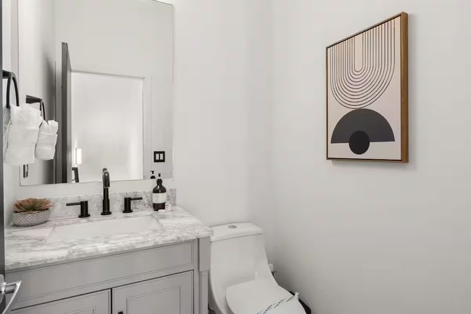 Bright modern powder room with marble countertop, matte black faucet, white toilet, small succulent accent, and framed geometric wall art.