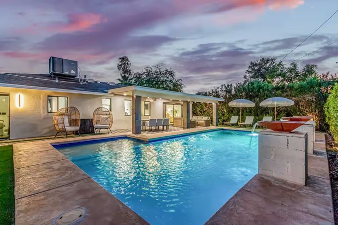 Twilight suburban backyard with a sparkling rectangular swimming pool, covered patio dining and lounge area, rattan egg chairs, sun loungers with umbrellas and surrounding greenery.