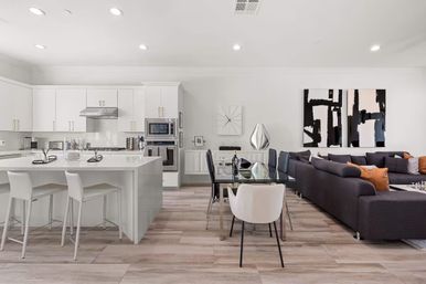 Bright open-concept modern kitchen and living room with a white island and bar stools, stainless-steel appliances, glass dining table, dark gray sectional sofa with orange accent pillows, abstract black-and-white wall art, and light wood-look flooring.