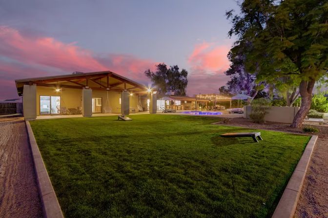Sunset backyard scene with a manicured lawn, covered pavilion with hanging chairs, illuminated pool, string lights and cornhole boards