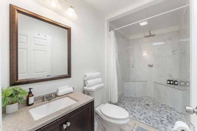 Bright modern home bathroom with dark wood vanity and granite countertop, framed mirror, stacked white towels, potted plant, and a large walk-in shower with marble-look wall tiles, pebble floor and rainfall showerhead.