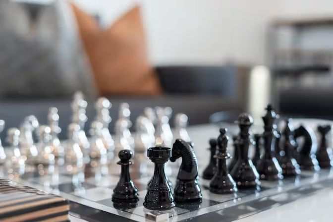 Close-up of glossy black and clear glass chess pieces on a glass chessboard on a living room coffee table with a blurred sofa and throw pillows in the background.