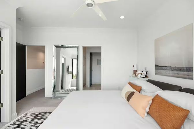 Bright modern coastal-style bedroom with white walls, king bed in white linens, orange accent pillows, large beach wall art above headboard, ceiling fan, full-length mirror by the doorway and minimalist decor