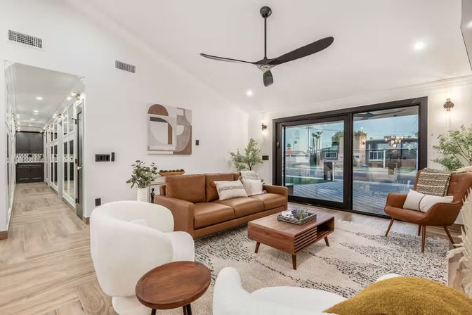 Bright modern living room with brown leather sofa, mid-century wood coffee table, white swivel chairs, large black-framed sliding glass doors overlooking a palm-lined suburban street, ceiling fan, indoor plants and neutral area rug.