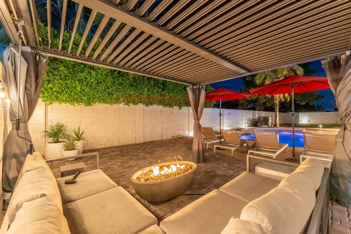 Cozy modern backyard patio at dusk with a slatted pergola, cushioned lounge sofa around a lit stone fire pit, brick pavers, red poolside umbrellas and loungers beside an illuminated swimming pool framed by hedges and palm trees.