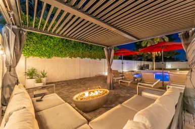 Cozy modern backyard patio at dusk with a slatted pergola, cushioned lounge sofa around a lit stone fire pit, brick pavers, red poolside umbrellas and loungers beside an illuminated swimming pool framed by hedges and palm trees.
