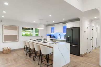 Bright modern open-concept kitchen in a sunny suburban home with a marble waterfall island and five bar stools, white cabinetry, black French-door fridge, blue LED under-cabinet lighting and wood-look plank floors.