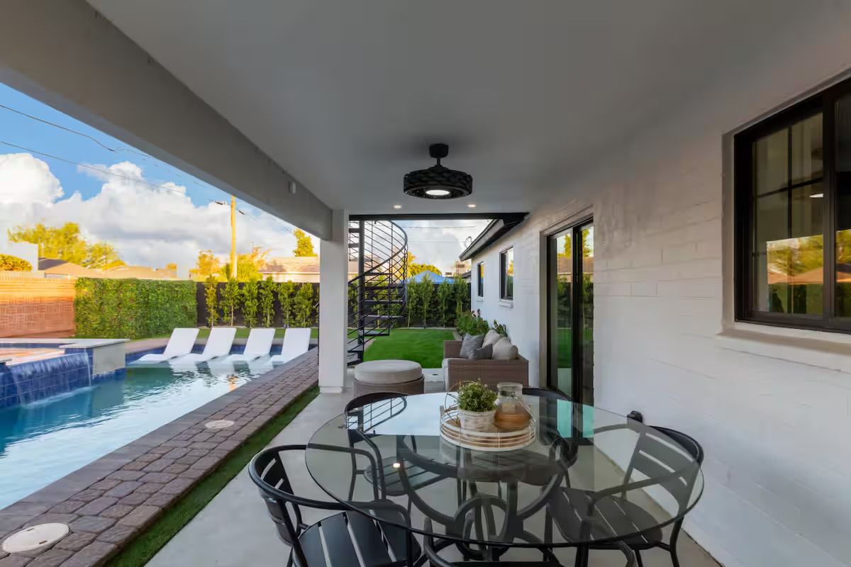 Covered modern backyard patio with glass dining table and chairs, cozy outdoor sofa, spiral staircase, and a pool with waterfall and in-pool loungers