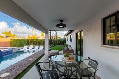 Covered modern backyard patio with glass dining table and chairs, cozy outdoor sofa, spiral staircase, and a pool with waterfall and in-pool loungers