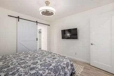 Bright modern bedroom interior with black-and-white floral duvet, wall-mounted flat-screen TV, white sliding barn door closet, circular ceiling light/fan, and light wood floors — cozy minimalist design.
