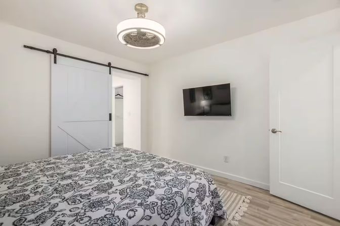 Bright modern bedroom interior with black-and-white floral duvet, wall-mounted flat-screen TV, white sliding barn door closet, circular ceiling light/fan, and light wood floors — cozy minimalist design.