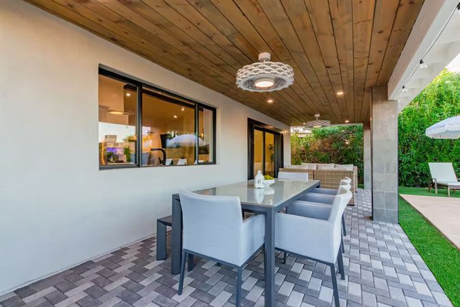 Inviting covered backyard patio with a modern glass-top dining table and cushioned chairs under a wood-plank ceiling with pendant lights, paver flooring and lush hedge backdrop.