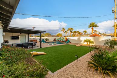 Sunny desert backyard with covered patio and outdoor seating, artificial turf lawn, pool with a colorful mural, palm trees and cornhole boards