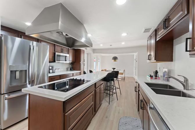 Bright modern kitchen with long white quartz island and induction cooktop under a stainless steel range hood, dark wood cabinets, stainless refrigerator and dishwasher, breakfast bar with black stools, pendant light over dining area, and light hardwood floors.