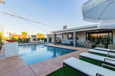 Modern suburban backyard pool and patio with rectangular swimming pool, cushioned lounge chairs under a fringed umbrella, covered outdoor dining and seating area, and a fireplace across the pool.