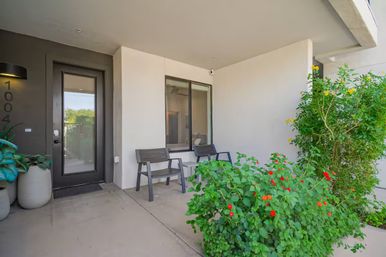 Contemporary apartment entrance with a black glass door, cozy covered front patio featuring two gray chairs and a small table, potted greenery and lush flowering shrubs with red and yellow blooms.