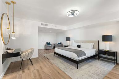 Spacious modern king bedroom suite with platform bed, gray throw and pillows, black nightstands with lamps, comfy seating area, wall mirror and pendant lights, light wood floors and neutral decor.