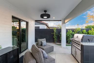 Covered backyard patio with gray modular lounge chairs and cushions, stainless steel gas grill, black privacy wall, sliding glass doors, modern overhead light, and green shrubs under a blue sky.