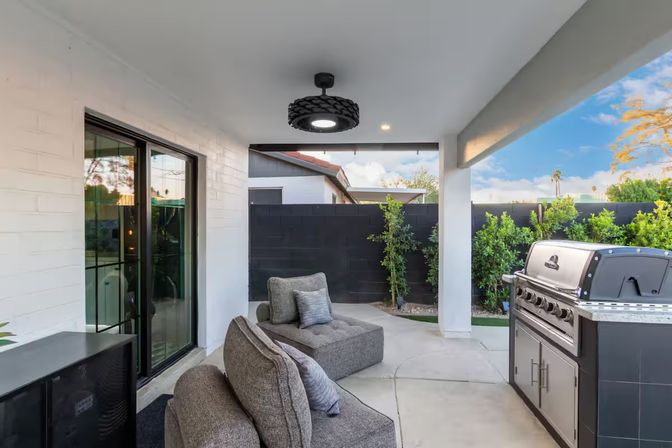 Covered backyard patio with gray modular lounge chairs and cushions, stainless steel gas grill, black privacy wall, sliding glass doors, modern overhead light, and green shrubs under a blue sky.