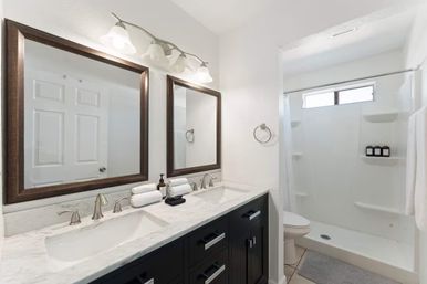 Bright spa-like modern bathroom with marble double-sink countertop, dark wood vanity, two framed mirrors, brushed nickel faucets, and a white walk-in shower with built-in shelves and toiletries.