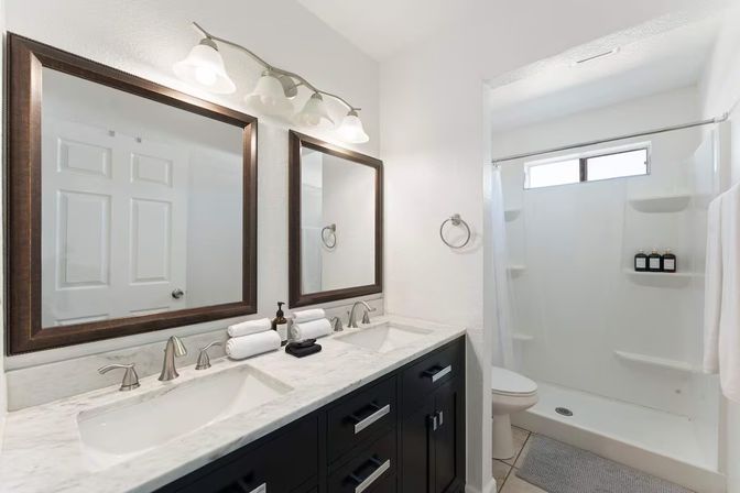 Bright spa-like modern bathroom with marble double-sink countertop, dark wood vanity, two framed mirrors, brushed nickel faucets, and a white walk-in shower with built-in shelves and toiletries.