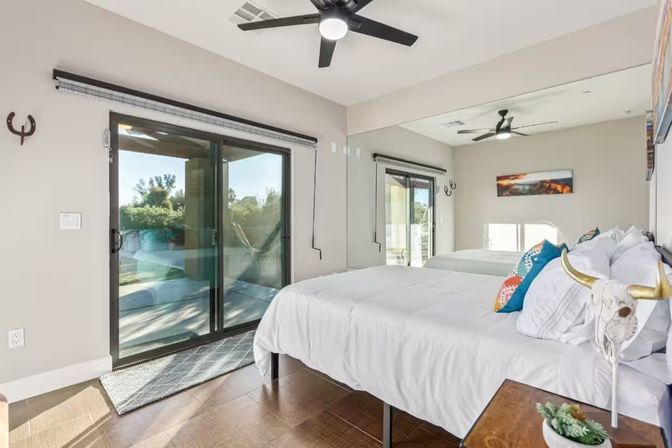 Modern bedroom with white bedding and colorful pillows, mirrored wall, black ceiling fan, wooden floor and sliding glass doors opening to a sunny backyard patio.