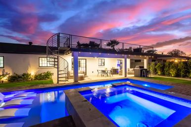 Modern backyard at sunset: blue LED-lit pool and raised spa, rooftop deck with spiral staircase, covered patio dining and lounge under colorful sky