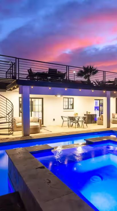 Modern backyard at sunset: blue LED-lit pool and raised spa, rooftop deck with spiral staircase, covered patio dining and lounge under colorful sky