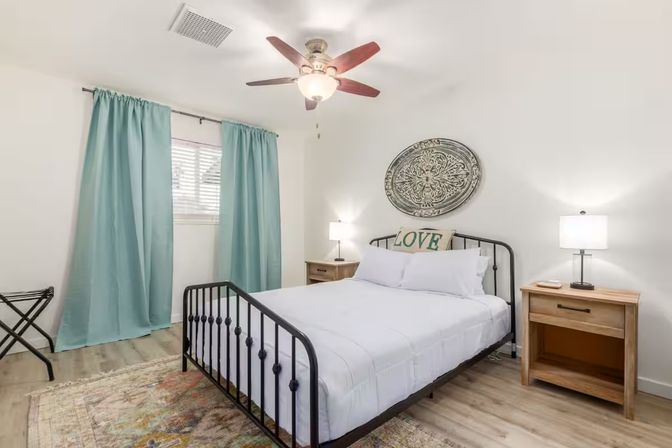 Cozy bright bedroom with black metal bed frame, white bedding and a "LOVE" pillow, teal curtains over the window, wooden nightstands with lamps, decorative round wall medallion, ceiling fan, light hardwood floors and a colorful area rug.