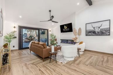 Bright, cozy-chic living room with a brown leather sofa and two white swivel chairs around a wooden coffee table, wall-mounted TV above a linear fireplace, large sliding glass doors, wood-look plank flooring, ceiling fan, plants and a framed black-and-white longhorn print.
