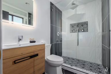 Modern bathroom with glass walk-in shower, black-and-white geometric floor tiles, vertical dark tile accent, wooden vanity with illuminated mirror and white toilet