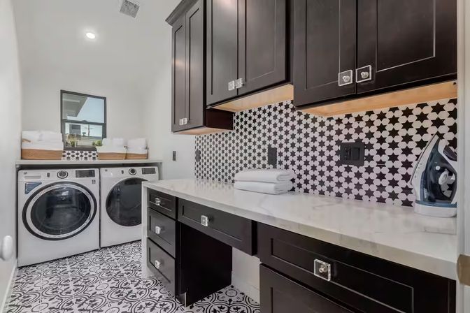 Stylish modern laundry room with front-loading washer and dryer, dark shaker cabinets, white countertop, black-and-white star tile backsplash, patterned floor tiles, folding area and iron.