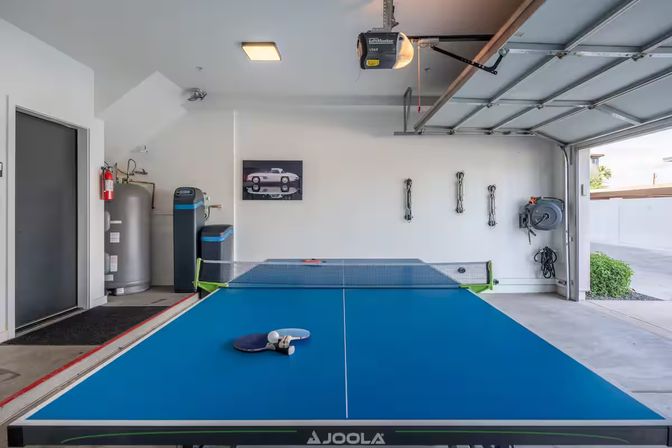 Blue table tennis (ping-pong) table with paddles and ball ready for a match in a bright residential garage recreation area, open garage door, wall-mounted hose reel, water tank and fire extinguisher visible.