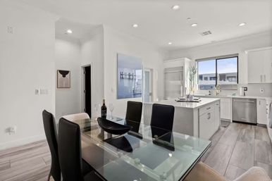 Bright, airy modern open-concept kitchen and dining area with glass-top table and black chairs, white island with marble countertop, stainless-steel appliances and large window