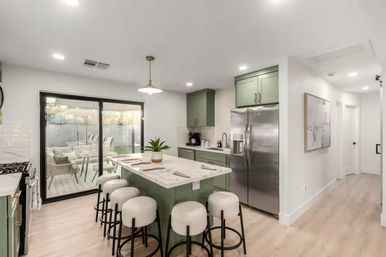 Bright modern open-concept kitchen with sage-green cabinets, marble-topped island with six upholstered bar stools, stainless-steel refrigerator, pendant light, light wood floors, and large sliding glass doors to a patio.