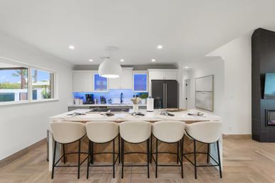Bright modern open-concept kitchen with large white island and five cushioned bar stools, pendant light, stainless appliances, blue-lit backsplash, and sunlit window showing palm trees — stylish space for entertaining.