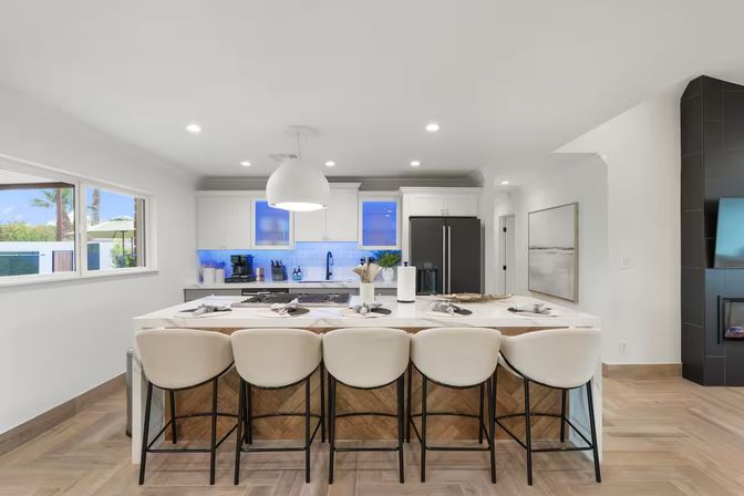 Bright modern open-concept kitchen with large white island and five cushioned bar stools, pendant light, stainless appliances, blue-lit backsplash, and sunlit window showing palm trees — stylish space for entertaining.