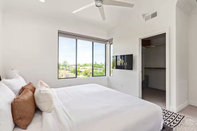 Sunlit modern bedroom with white king bed and brown accent pillows, ceiling fan, wall-mounted TV and large window overlooking suburban greenery and palm trees.