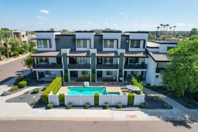 Modern three-story townhouse complex with white-and-gray facade and balconies overlooking a fenced turquoise communal pool, lounge chairs and striped umbrella, landscaped hedges and palm-lined residential street on a sunny day.