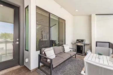 Modern covered balcony patio with gray outdoor sofa and pillows, gas grill, white slatted dining table and potted succulent by glass door and windows