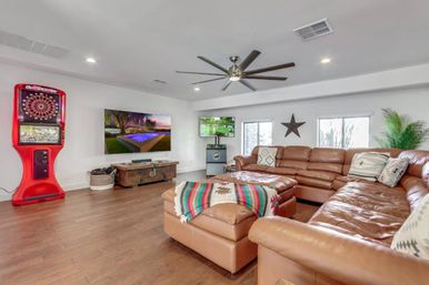 Spacious bright home entertainment room with a large brown leather sectional and ottoman draped in a colorful blanket, wall-mounted TV above a rustic wooden chest, red electronic dartboard arcade, ceiling fan, hardwood floors, star wall decor and potted plant.