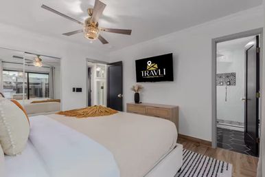 Bright modern vacation-rental bedroom with neutral bedding and mustard throw, mirrored sliding closet, ceiling fan, wall-mounted flat-screen TV, wooden dresser and doorway to tiled bathroom.