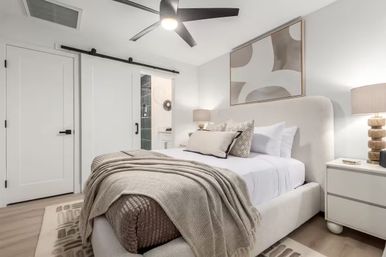 Cozy modern neutral bedroom with upholstered bed, soft layered throws and pillows, abstract wall art, ceiling fan, barn-style sliding door to a tiled en-suite bathroom, and matching nightstands with lamps.