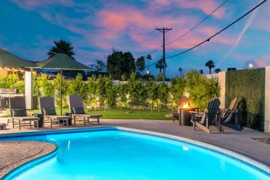 Dusk backyard pool with glowing blue water, lounge chairs and umbrellas, a lit fire pit, palm trees and a pink sunset sky.