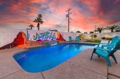 Backyard pool at sunset with turquoise Adirondack chairs, small waterfall feature and a vibrant mural reading Scottsdale on a white block wall, palm trees and pink-orange sky