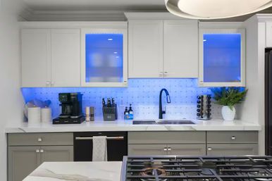Sleek modern white kitchen with blue LED under-cabinet lighting, marble countertops, black pull-down faucet, coffee maker, knife block, potted fern, and gas cooktop in foreground.
